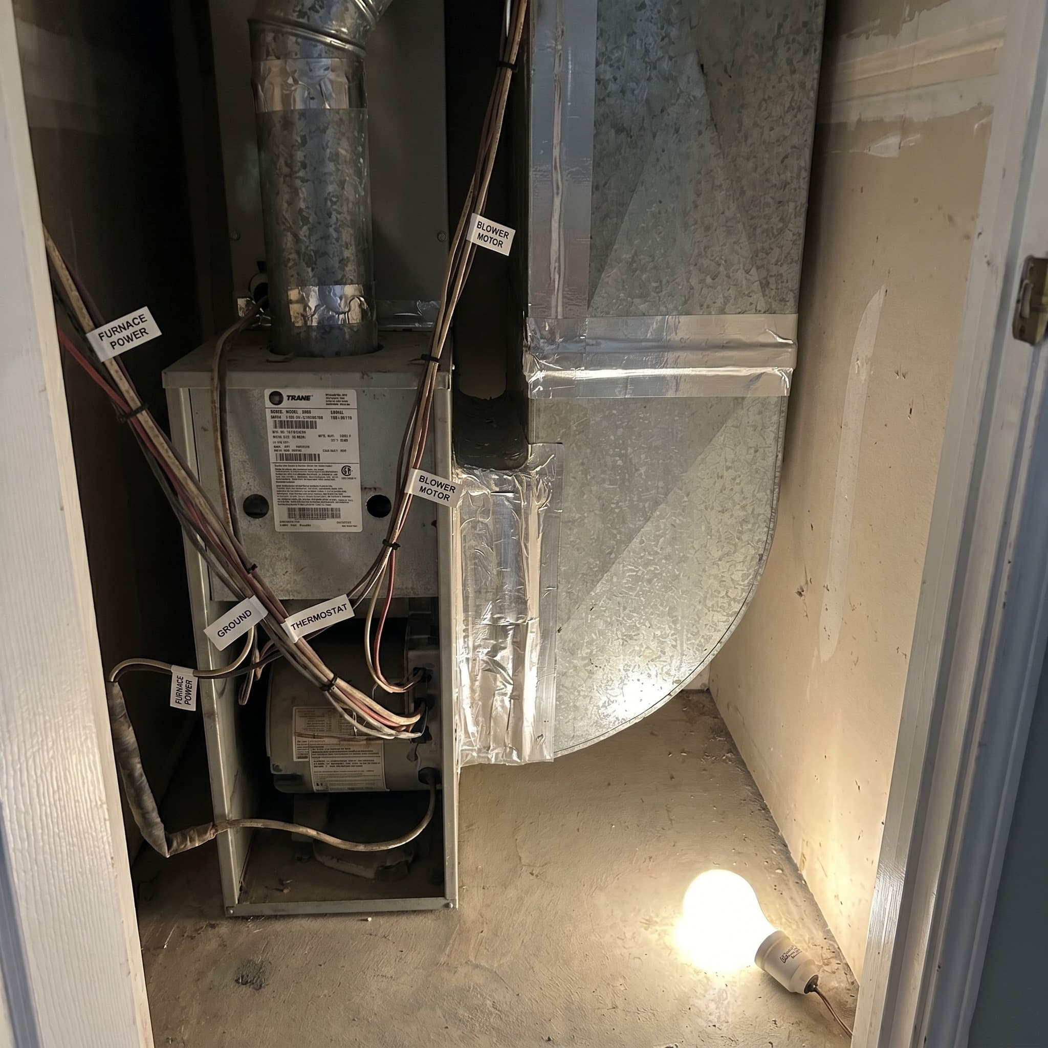 Residential air handler closet interior — neatly labeled wire bundles with printed tags, clean wrapped ductwork seams, model-and-serial sticker prominent on the unit