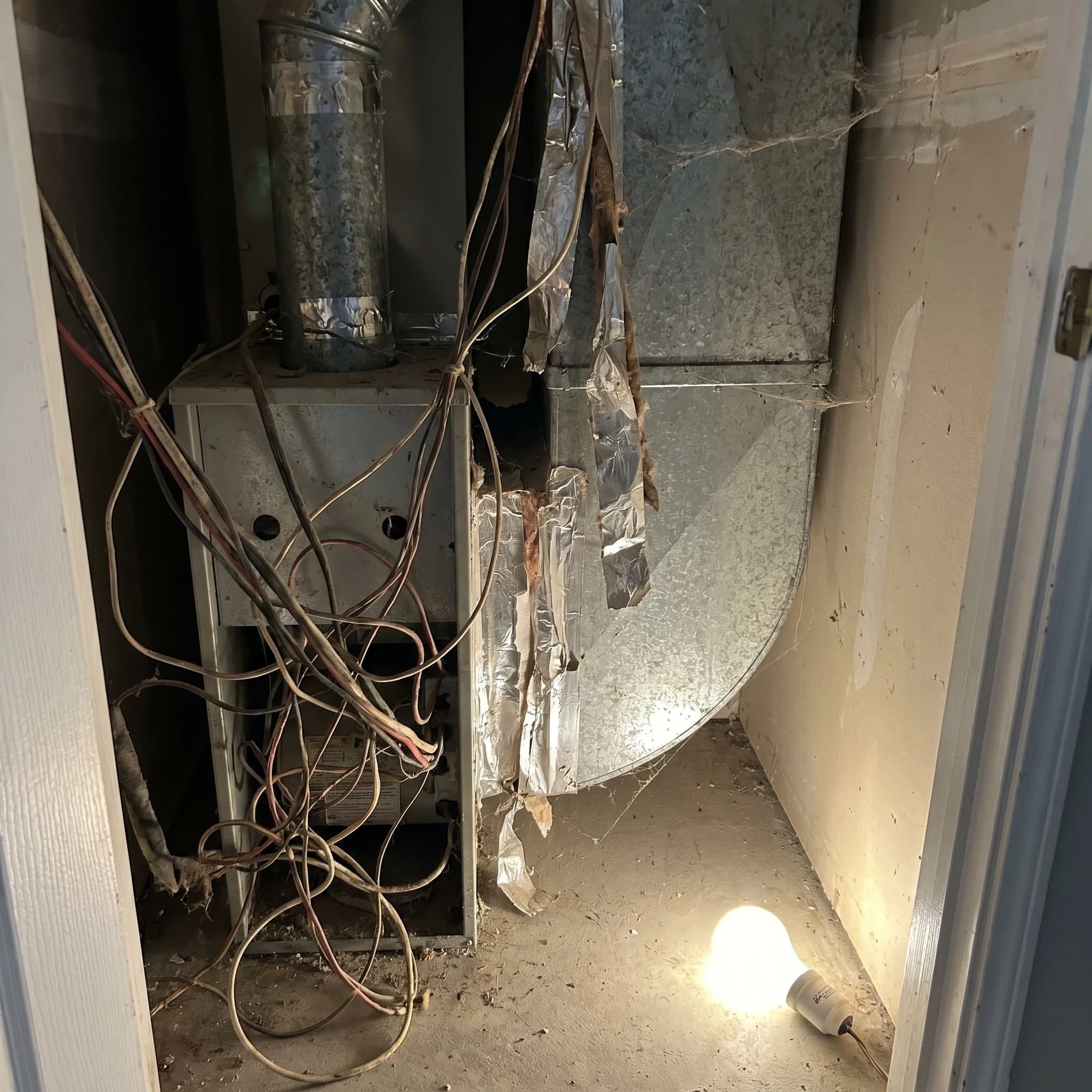 Residential air handler closet interior — exposed wires running in disorganized bundles with no labels and dusty unwrapped ductwork seams