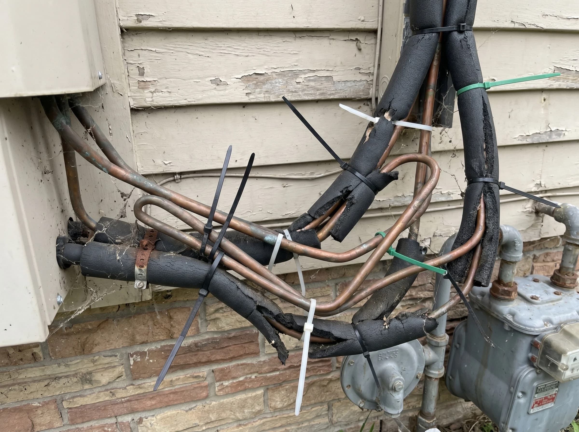 Tangled refrigerant line install on a residential exterior wall — kinked copper, loose insulation wrap, mismatched zip-tie support